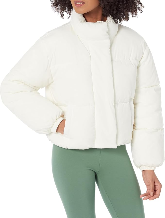 A woman wearing a crisp white, cropped full-zip puffer jacket with a high stand-up collar, quilted paneling, and elasticized cuffs, paired with sage green leggings