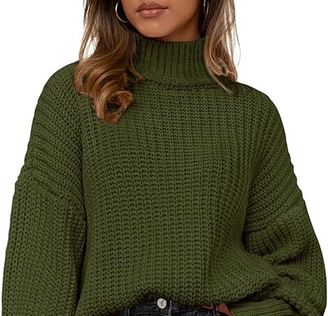 A woman wearing an oversized, olive green chunky knit turtleneck sweater with long, voluminous sleeves, partially tucked into dark grey jeans.