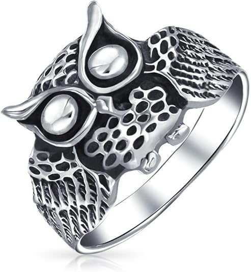 A silver-toned wide band ring featuring a detailed owl face as the centerpiece. The owl has large, prominent eyes and textured feathers, with the band itself mimicking the appearance of layered wings or feathers with an antiqued finish.