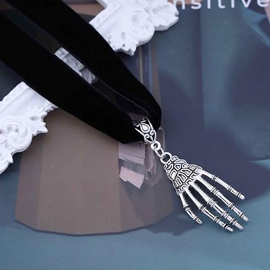 A close-up of a gothic choker necklace featuring a thick black velvet band. It is adorned with a detailed silver skeleton hand pendant dangling from a decorative silver bail. The piece is displayed on a textured surface next to a white vintage-style frame.