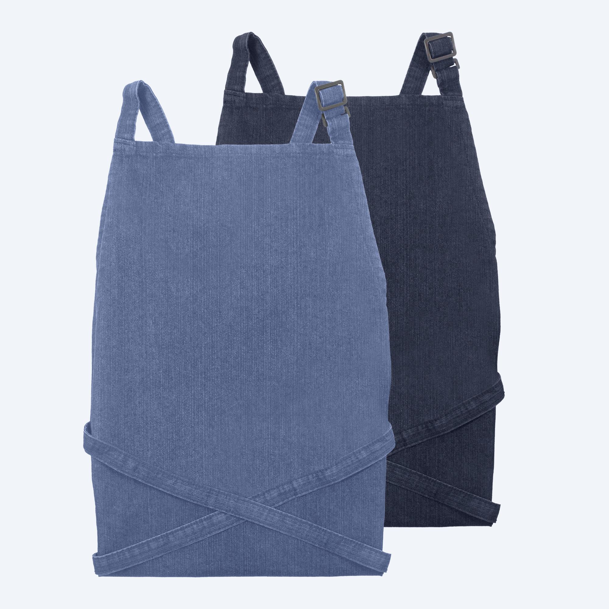 Two folded, premium recycled denim bib aprons—one in a light blue wash and one in deep indigo—featuring metal adjustment buckles on the neck straps and cross-back style waist ties.