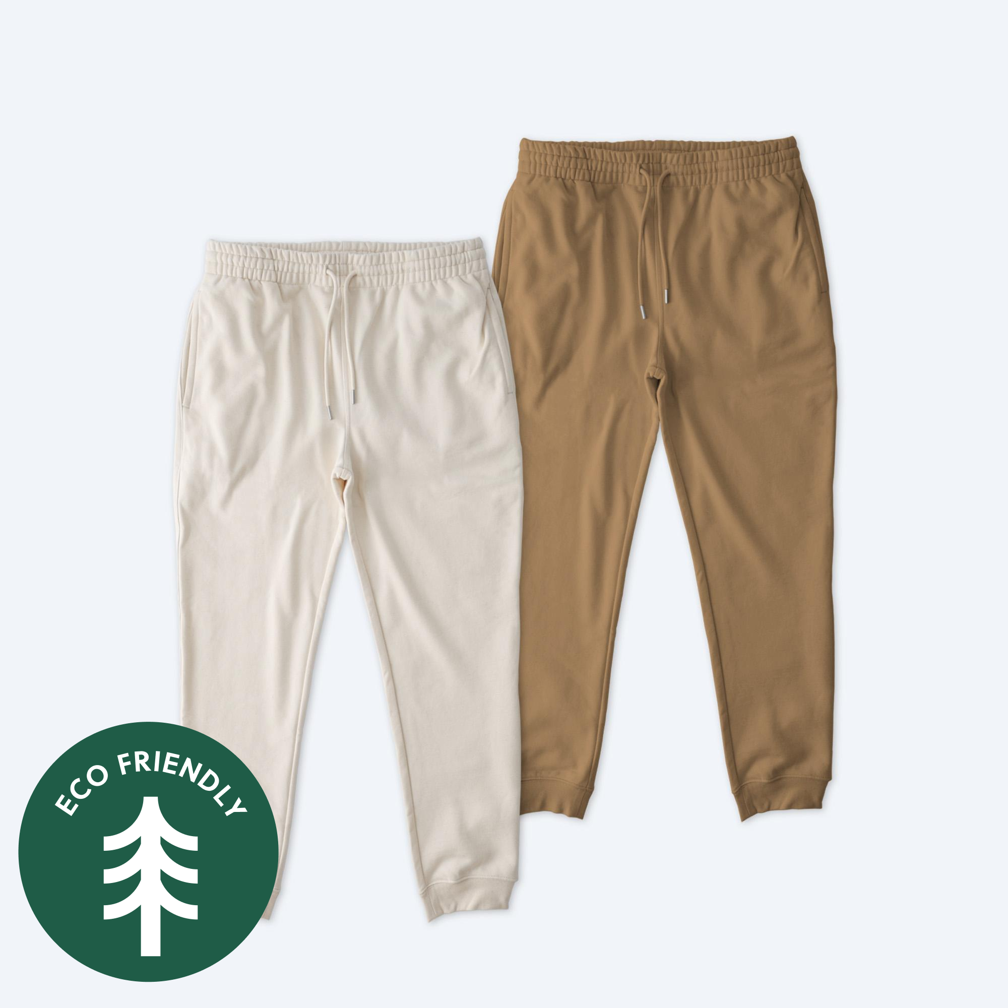 Two pairs of Stanley Stella Unisex Flyer Joggers in Natural Raw and Desert Dust colors, featuring a green "Eco Friendly" tree logo, elastic waistbands with drawstrings, and ribbed cuffs.